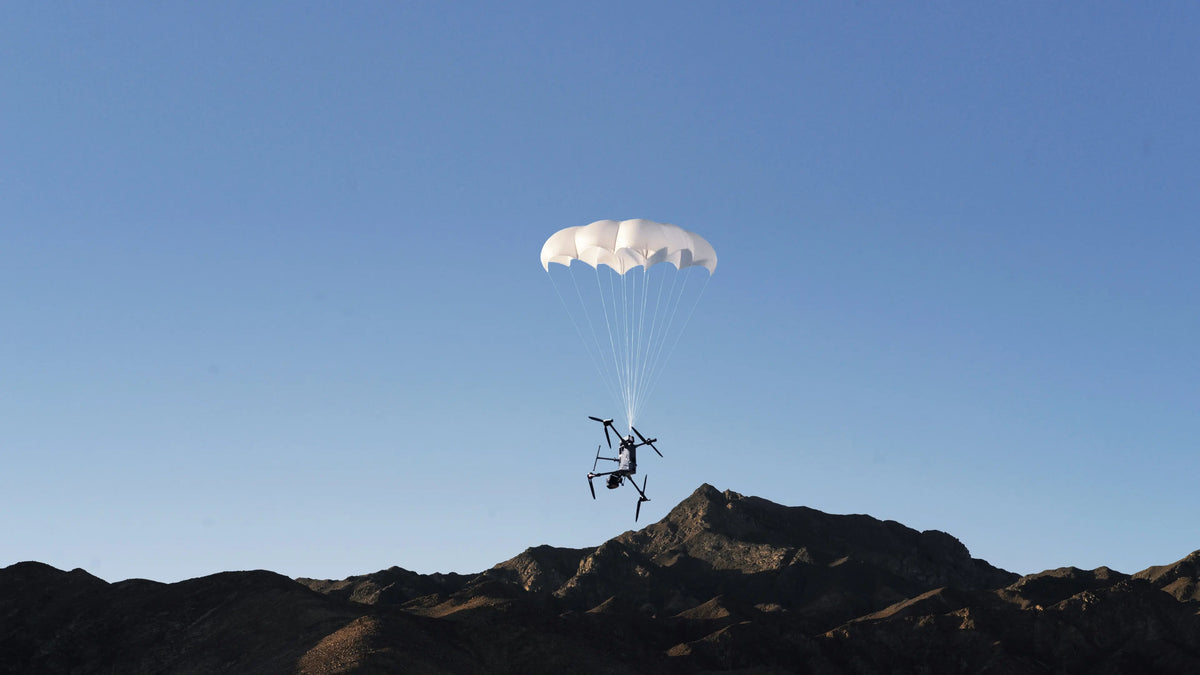 DJI Matrice 400 descending under a deployed DJI AP100 Parachute canopy over mountain terrain
